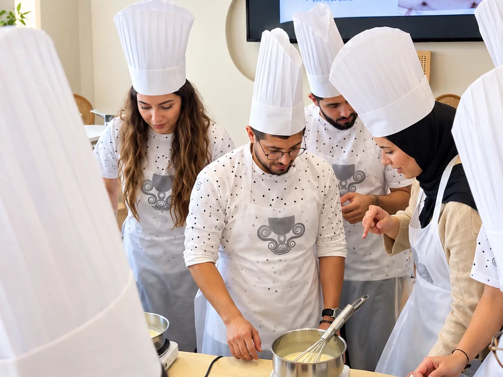 Une équipe en toque prépare une recette autour de casseroles, la fiche recette projetée derrière.